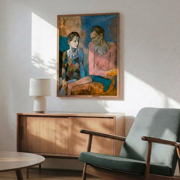 May include: Framed reproduction of Picasso's 1905 painting showing two circus performers - a young boy in diamond-patterned harlequin costume and seated adult figure in pink garment with white ruff collar. Artwork displays muted blues, pinks, and earth tones in classical style. Print hangs above mid-century modern wooden credenza with ceramic table lamp, positioned next to sage green upholstered armchair with wooden frame in bright, naturally-lit room.