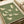 Japanese cranes in flight pattern on sage green textured background. White and cream colored birds with long necks and outstretched wings shown in various flying poses. Art print displayed on white paper with cream ribbon nearby, photographed from above on light wooden surface.