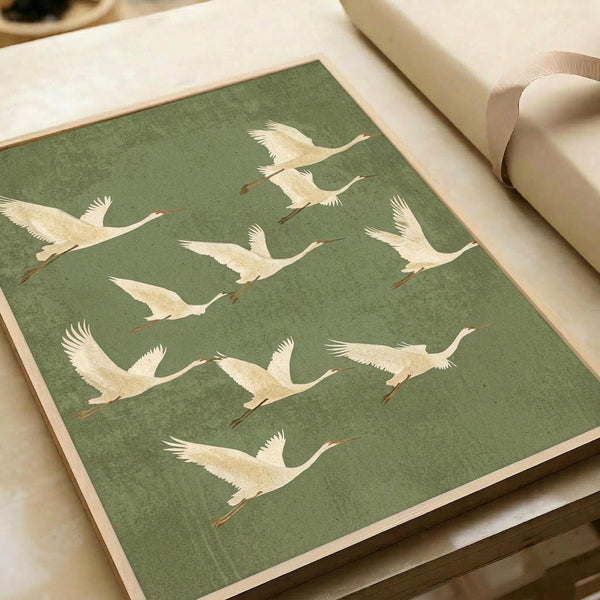 Japanese cranes in flight pattern on sage green textured background. White and cream colored birds with long necks and outstretched wings shown in various flying poses. Art print displayed on white paper with cream ribbon nearby, photographed from above on light wooden surface.