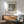 May include: Framed Monet Water Lilies poster print hanging above wooden credenza with table lamp, featuring impressionist pond scene with floating lilies in soft greens, blues and purples. Mid-century modern room setting with sage green upholstered chair, round coffee table, and natural sunlight casting geometric shadows on white wall.