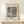May include: Framed art print of Monet's Water Lilies painting featuring impressionist brushstrokes in soft blues, greens, and purples depicting lily pads floating on reflective pond water. Print displays "MONET" at top and "Water Lilies" at bottom in elegant typography. Light wood frame sits on pale wooden shelf beside small white wrapped gift with evergreen sprig against cream wall with natural lighting.
