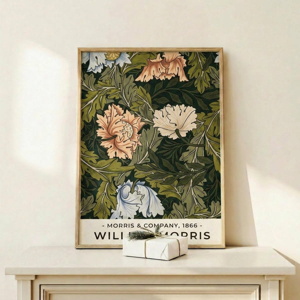 May include: Framed botanical poster with William Morris-style floral pattern featuring pink, white and coral flowers with lush green foliage on dark background, displayed on light wood shelf beside small wrapped gift with evergreen sprig against cream wall