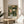 May include: Framed botanical art print displayed on round wooden side table next to beige upholstered chair. Print features vintage floral pattern with coral, white and blue blooms among dense green foliage in decorative Arts and Crafts style. Natural wood frame complements warm sunlit room with neutral walls and plant shadows visible.