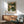May include: Framed botanical poster with green leafy pattern and peach flowers displayed above wooden sideboard in modern living room. Mid-century green upholstered chair with wooden arms positioned nearby. White ceramic table lamp on credenza. Bright natural sunlight casting shadows across white wall and wooden furniture.