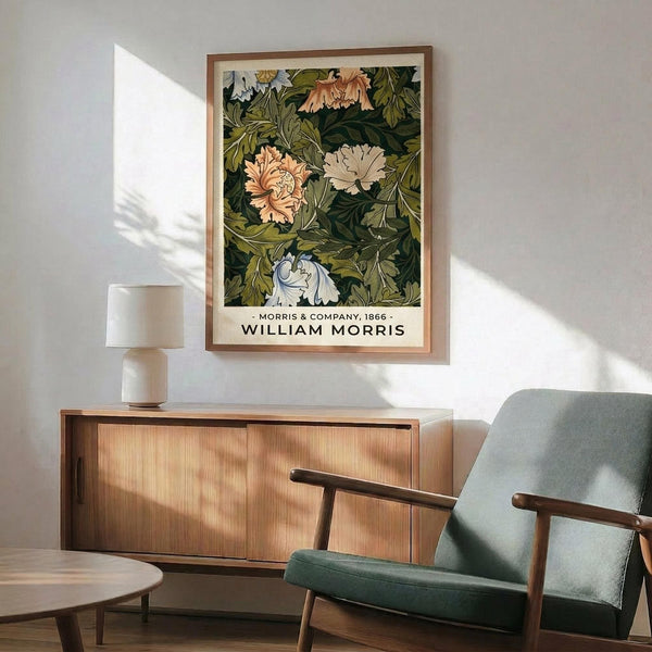 May include: Framed botanical poster with green leafy pattern and peach flowers displayed above wooden sideboard in modern living room. Mid-century green upholstered chair with wooden arms positioned nearby. White ceramic table lamp on credenza. Bright natural sunlight casting shadows across white wall and wooden furniture.