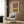 May include: Abstract geometric poster with terra cotta and mustard yellow color blocks featuring black line arch designs, displayed in light wood frame on warm brown wood paneled wall above cream upholstered armchair with geometric patterned pillow, brass floor lamp and side table with open book nearby