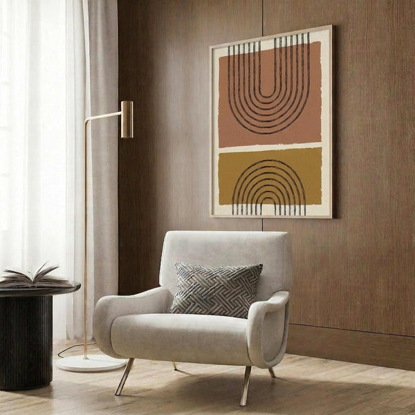 May include: Abstract geometric poster with terra cotta and mustard yellow color blocks featuring black line arch designs, displayed in light wood frame on warm brown wood paneled wall above cream upholstered armchair with geometric patterned pillow, brass floor lamp and side table with open book nearby
