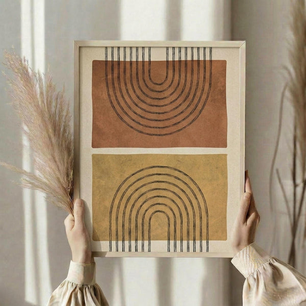 May include: Person in beige blouse holding framed abstract art print featuring two rainbow arch designs - one terracotta orange arch extending upward and one mustard yellow arch extending downward with connecting vertical lines, set against neutral backgrounds in light wood frame, with dried pampas grass visible to the left