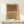 May include: A framed abstract art print featuring two arched rainbow shapes in terracotta orange and mustard yellow tones with black line details, displayed on a light wood console table next to a small wrapped gift with greenery. The artwork is set in a natural wood frame against a cream colored wall in minimalist home decor styling.