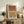 May include: Modern abstract art print featuring two rainbow arch shapes in terracotta orange and mustard yellow with black line details, displayed in light wood frame on rustic wooden sideboard. Room scene shows neutral decor with fern plant, ceramic vase, stacked books, and beige chair with brown leather pillow in warm natural lighting.