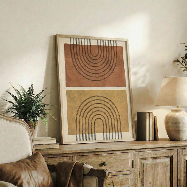 May include: Modern abstract art print featuring two rainbow arch shapes in terracotta orange and mustard yellow with black line details, displayed in light wood frame on rustic wooden sideboard. Room scene shows neutral decor with fern plant, ceramic vase, stacked books, and beige chair with brown leather pillow in warm natural lighting.
