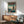 May include: Framed poster showing a lone figure in space suit standing on futuristic building rooftop overlooking neon-lit cyberpunk cityscape in teal and orange tones. Displayed above mid-century modern wooden credenza with white ceramic lamp, next to sage green upholstered armchair with wooden frame in bright, naturally lit room with white walls.