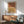 May include: Framed artwork with two abstract arches in terracotta and mustard yellow tones hanging above a wooden credenza. Modern living room setting features a sage green mid-century armchair, white ceramic table lamp, and round coffee table. Warm sunlight streams across white walls creating dramatic shadows.