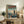May include: Framed poster showing a futuristic pilot in armor standing on a rooftop overlooking a sprawling cyberpunk cityscape with tall buildings and teal-orange sky. The artwork features Japanese text at the top and "LONE PILOT OF NEO-TOKYO" subtitle. Light wood frame rests on rustic wooden sideboard next to potted fern, books, ceramic vase, and brown leather chair in cozy living room setting with white walls.