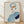 May include: Framed abstract portrait print leaning against white textured wall on light wood floor. Artwork shows stylized figure with blue-gray hair, orange and white face sections, purple lips, and blue clothing with dark navy accent. Natural wood frame, soft window light creates diagonal shadows on wall.
