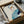 Framed abstract portrait artwork being unpacked from cardboard shipping box with protective tissue paper. The minimalist illustration features a stylized face with blue-gray hair, orange and purple facial features, and turquoise clothing against a cream background. Black frame sits in brown cardboard box surrounded by white packing materials on light surface.