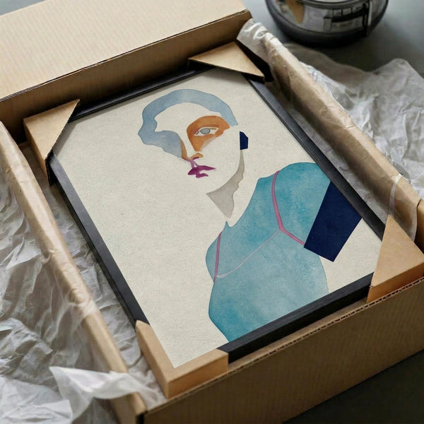 Framed abstract portrait artwork being unpacked from cardboard shipping box with protective tissue paper. The minimalist illustration features a stylized face with blue-gray hair, orange and purple facial features, and turquoise clothing against a cream background. Black frame sits in brown cardboard box surrounded by white packing materials on light surface.