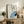 May include: Framed abstract portrait artwork in light wooden frame displayed on rustic wooden sideboard. Art features stylized face with blue hair, geometric color blocks in blues, orange, and purple on cream background. Scene includes fern plant in white pot, stacked books, ceramic vases, and beige upholstered furniture creating warm, modern living room setting.