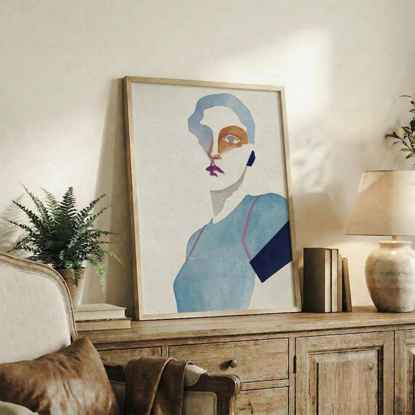 May include: Framed abstract portrait artwork in light wooden frame displayed on rustic wooden sideboard. Art features stylized face with blue hair, geometric color blocks in blues, orange, and purple on cream background. Scene includes fern plant in white pot, stacked books, ceramic vases, and beige upholstered furniture creating warm, modern living room setting.