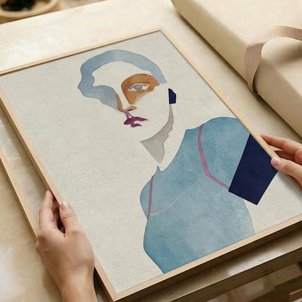 May include: Hands holding a framed minimalist portrait print featuring an abstract female figure in soft blue, navy, orange and purple tones. The artwork shows a stylized woman with flowing blue-gray hair wearing a light blue garment. The print is displayed in a thin wooden frame on cream wrapping paper with ribbon visible in the background.
