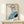 May include: Abstract minimalist portrait artwork in thin wooden frame leaning against white wall on light wood surface. Features stylized face with blue hair, orange eye area, purple lips, and geometric blue and navy clothing shapes on cream background. Small white wrapped gift with natural twine sits in front of frame.