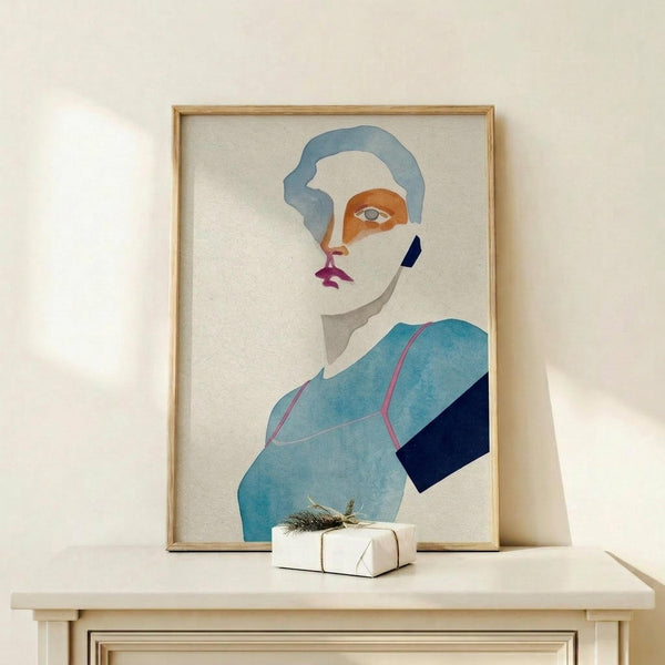 May include: Abstract minimalist portrait artwork in thin wooden frame leaning against white wall on light wood surface. Features stylized face with blue hair, orange eye area, purple lips, and geometric blue and navy clothing shapes on cream background. Small white wrapped gift with natural twine sits in front of frame.