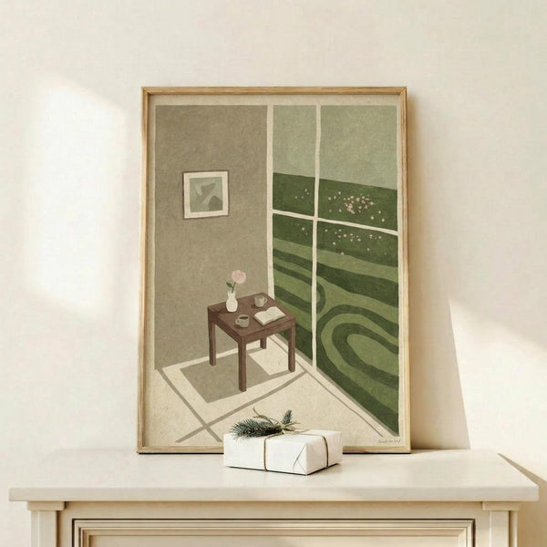 May include: Framed art print in light wood frame showing an interior room corner with window view of rolling green hills dotted with flowers. The room features sage green walls, a small dark wooden table with pink flower, open book and cup, plus a small framed artwork. A white wrapped gift with evergreen sprig sits on a light wooden surface below the frame against a cream wall.