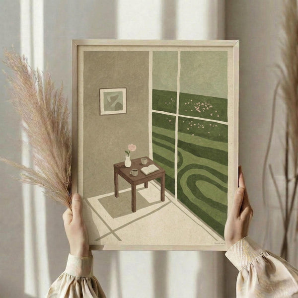May include: Hands holding a framed art print showing an interior room scene with sage green walls, white window frames overlooking rolling green hills with small flowers, a wooden side table with vase, book and cup, and a small framed artwork on the wall. Natural lighting with dried pampas grass visible to the left. Muted earth tones throughout with soft, illustrated style artwork.