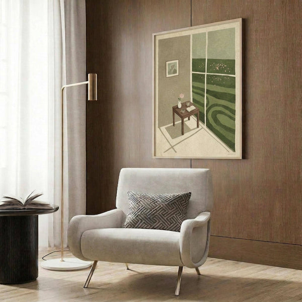 May include: Framed art print hanging on wood-paneled wall showing interior room scene with window view of green landscape, dark wooden table, and wall art. Modern beige armchair with geometric pillow sits below artwork. Gold floor lamp and dark side table with open book complete the styled room setting.