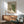 May include: Mid-century modern living room with sage green upholstered armchair and wood sideboard. Framed artwork on white wall depicts an interior scene with brown table, vase with pink flower, and window view of rolling green hills. Natural sunlight casts geometric shadows across the space. Contemporary table lamp with white shade sits on sideboard.