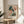 May include: Framed botanical art print featuring abstract painted flowers in soft pink, coral, green and white tones displayed on round wooden side table next to beige upholstered chair. Natural sunlight creates shadows on white wall behind the artwork, with palm plant leaves visible in corner of minimalist living room setting.