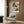 May include: Light beige armchair with geometric pillow positioned against wood-paneled wall. Above hangs framed botanical art print featuring abstract floral brushstrokes in soft pink, coral, green and cream tones. Gold floor lamp and dark round side table with open book visible nearby. Warm natural lighting creates cozy modern interior setting.
