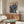May include: Framed poster featuring anime-style scene with soldier in green uniform facing large mecha robot in futuristic Neo Tokyo cityscape at night, displayed on wooden credenza in modern living room with white curved chair, brass floor lamp, oval glass coffee table, and soft natural lighting