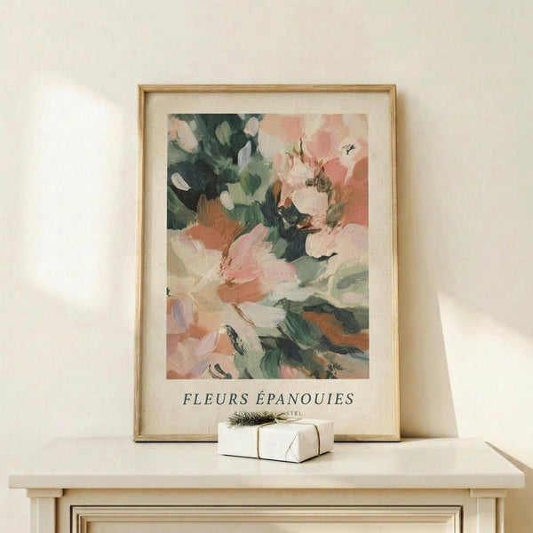 May include: Framed abstract floral art print in light wood frame, featuring soft brushstrokes in coral pink, sage green, and cream tones. Display shows artwork leaning against white wall on natural wood console table beside small white wrapped gift with green sprig. Warm natural lighting creates gentle shadows on minimalist neutral background.