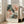 May include: A framed abstract floral art print leaning against a white wall next to a beige upholstered chair. The artwork features loose, painterly brushstrokes in soft pink, coral, sage green, and cream tones suggesting blooming flowers. The piece has French text at the bottom and is displayed in a light wood frame. Natural sunlight streams through nearby windows onto hardwood floors.