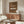 May include: Brown vintage-style poster featuring classic white Porsche 911 sports car with "911 CLASSIC" text and "1964" year marking, displayed in wooden frame above mid-century modern walnut credenza. Room shows white upholstered chair, brass floor lamp, white table lamp, glass-top coffee table, and neutral walls with natural lighting.