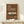 May include: Vintage-style brown poster featuring cream-colored Porsche 911 sports car with "PORSCHE 911 CLASSIC 1964" text in retro typography, displayed in natural wood frame on light wooden surface beside small wrapped white gift with pine sprig, photographed against cream wall with soft plant shadows