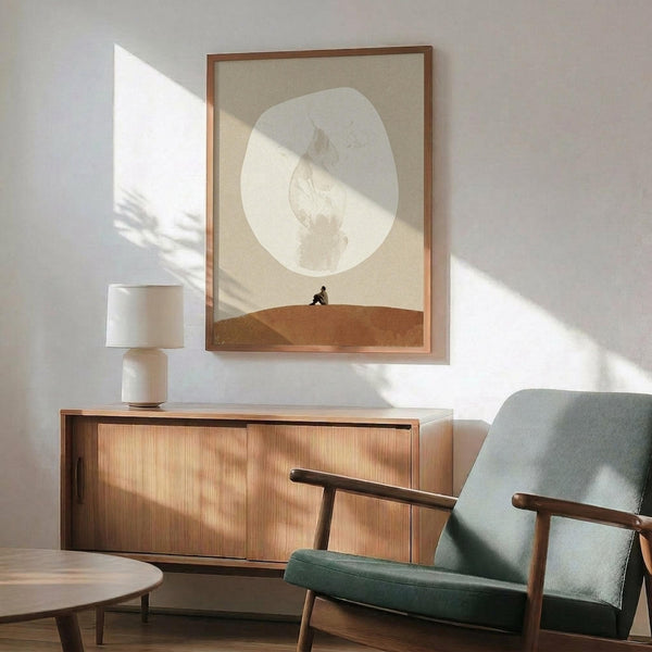 May include: Framed surrealist art print displayed above wooden sideboard in modern living room. Artwork shows small silhouetted figure sitting on sandy hill beneath large pale moon with botanical leaf motifs against neutral beige background. Natural sunlight creates dramatic shadows across white wall. Mid-century furniture includes teal upholstered armchair with wooden frame and ceramic table lamp.
