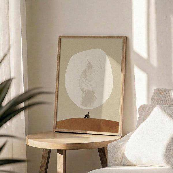 May include: Framed surrealist poster print displayed on round wooden side table next to beige upholstered chair. Artwork features minimalist desert landscape with large pale moon containing textured leaf pattern, small silhouetted figure on orange sand dune against cream background. Natural lighting creates soft shadows across neutral-toned room setting with plant leaves visible in corner.