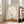 May include: Framed minimalist poster leaning against beige sofa in bright living room with large windows. Artwork shows small silhouetted figure sitting on orange-brown landscape beneath oversized pale moon on cream background. Light wood frame, modern neutral decor setting.