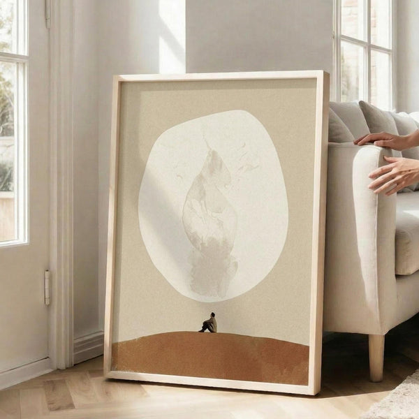 May include: Framed minimalist poster leaning against beige sofa in bright living room with large windows. Artwork shows small silhouetted figure sitting on orange-brown landscape beneath oversized pale moon on cream background. Light wood frame, modern neutral decor setting.