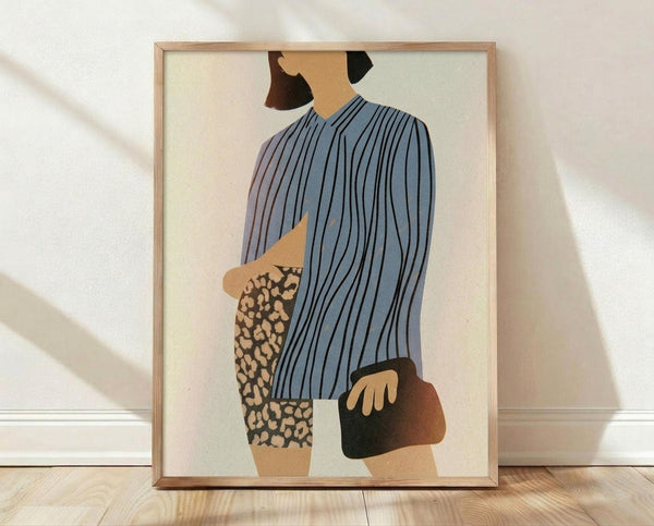 May include: Framed poster print leaning against white textured wall on light wood floor with natural shadows. Shows minimalist illustration of figure from behind wearing blue striped top, holding brown handbag, with leopard print pants. Artwork has cream background in natural wood frame.