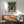 May include: Framed poster showing a silhouetted figure standing in a rainy cyberpunk alley with a large mech robot looming overhead, featuring teal and orange neon colors with Japanese text below. The artwork is displayed above a mid-century modern wooden credenza with a white table lamp, next to a mint green upholstered armchair in a bright, minimalist living room with white walls and natural sunlight streaming across the space.