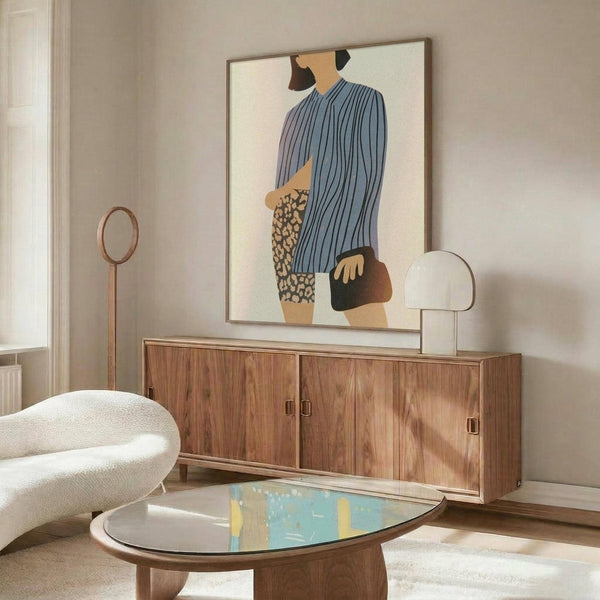 May include: Modern living room with abstract artwork on wall showing figure in blue striped top and leopard print shorts carrying brown bag. Wood credenza below with glass table lamp. White curved chair and oval coffee table with colorful glass top in foreground. Warm neutral walls and natural lighting.