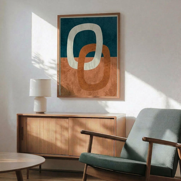 May include: Abstract modern poster with interlocking oval rings in cream, terracotta orange, and navy blue colors, displayed in wooden frame above mid-century credenza. Scene shows mint green upholstered armchair with wooden arms, white ceramic table lamp, and warm natural lighting creating shadows on white wall.