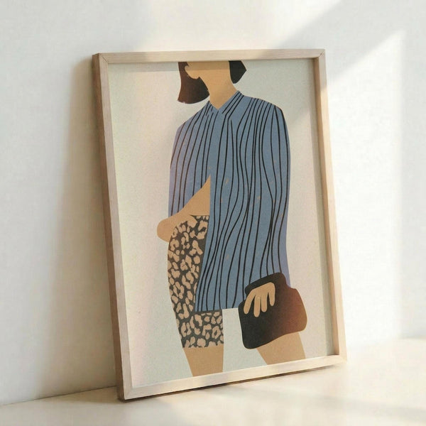 Framed abstract fashion illustration showing a stylized female figure wearing a blue and white striped top and leopard print skirt, holding a dark handbag. The minimalist artwork features geometric shapes in muted tones of blue, beige, and brown against a cream background. The print is displayed in a light wooden frame, leaning against a white wall on what appears to be a white surface.