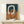 May include: Abstract art print in light wood frame showing interlocking oval rings in cream, terracotta orange, and navy blue with textured watercolor-style finish. Print displayed on light wood shelf with small wrapped gift box decorated with evergreen sprig against cream wall background.