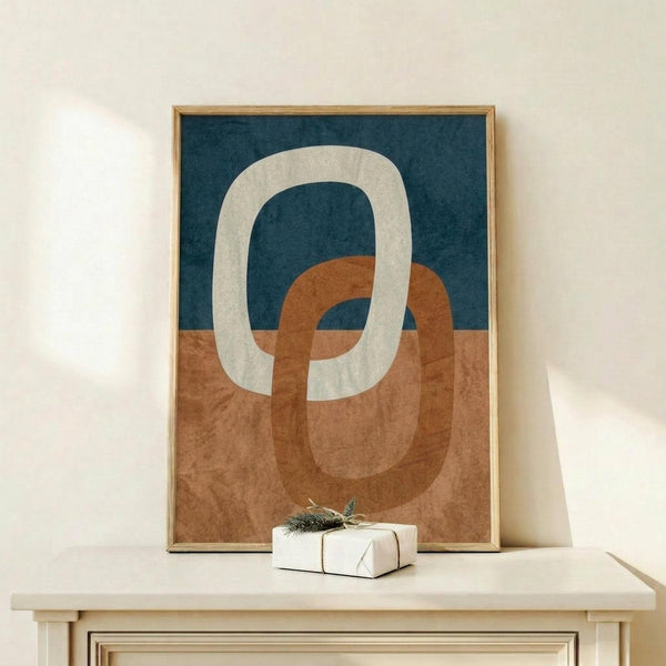 May include: Abstract art print in light wood frame showing interlocking oval rings in cream, terracotta orange, and navy blue with textured watercolor-style finish. Print displayed on light wood shelf with small wrapped gift box decorated with evergreen sprig against cream wall background.