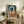 May include: Abstract geometric artwork with interlocking oval and circular shapes in terracotta orange, navy blue, and cream colors, displayed in a light wood frame on a rustic wooden sideboard. The framed print sits alongside potted plants, ceramic vases, books, and a cozy living room setting with neutral walls and a brown leather chair visible in the foreground.
