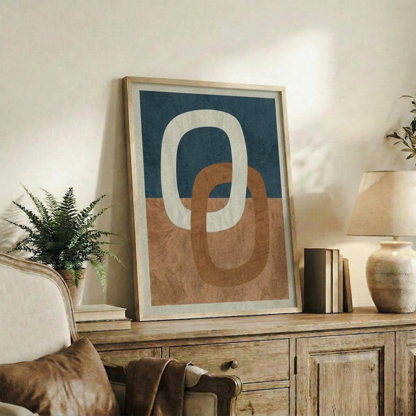 May include: Abstract geometric artwork with interlocking oval and circular shapes in terracotta orange, navy blue, and cream colors, displayed in a light wood frame on a rustic wooden sideboard. The framed print sits alongside potted plants, ceramic vases, books, and a cozy living room setting with neutral walls and a brown leather chair visible in the foreground.